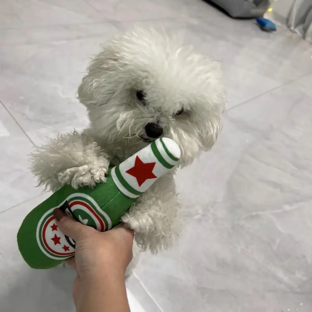 Small white fluffy puppy chewing on the green squeaky beer bottle plush toy